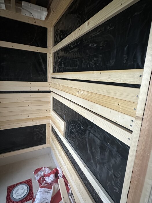 Bastubyggnation med träläkt och svart plast på väggar. Ventilationslucka i golv. Mattsäck och rördelar synliga på golvet.