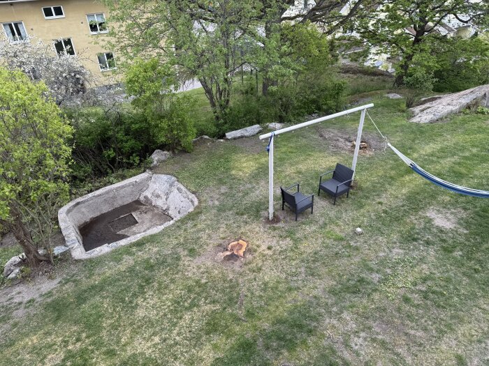 Trädgård med två stolar, en hängmatta och en sågstubbe på gräsmattan, omgiven av buskage och ett hus i bakgrunden.