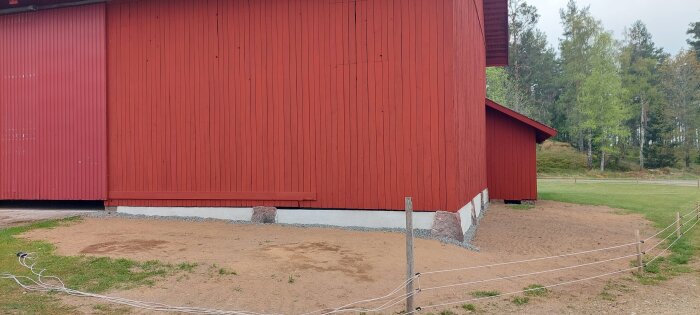 Röd byggnad med nypustad grund, omgiven av bar jord och väntande gräs.