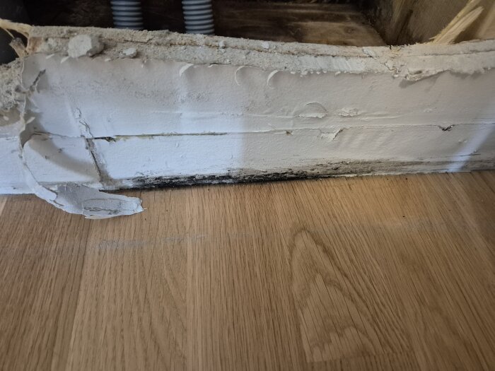 Vattenskada och fuktmärken vid nederkanten av en vit vägg över ett trägolv, med sprickor och avskavd färg nära golvsockeln.