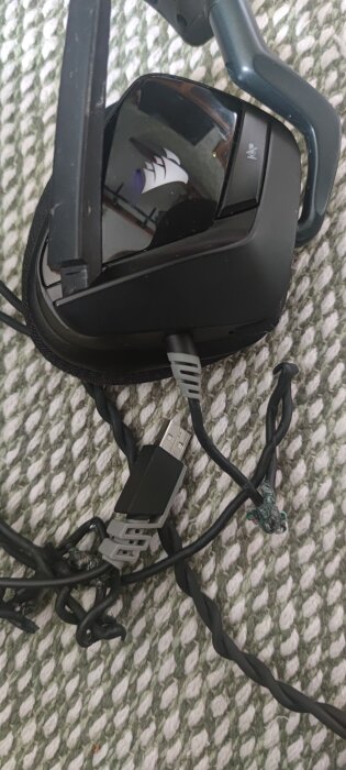 Svart gamingheadset med synlig USB-kabel och skadad kabel på en vit-grönrandig matta.