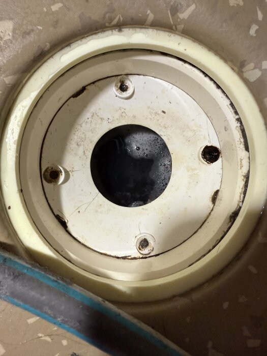 Vit rund konstruktion med skruvar runt kanten och en central öppning delvis fylld med vatten. Skruvarna ser åldrade ut.