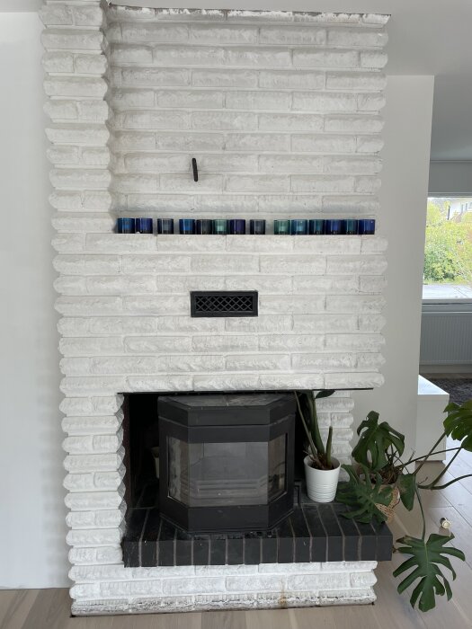 Vit tegelspis med mörka glaslyktor ovanpå och en svart kamin i öppningen, omgiven av krukväxter på golvet.