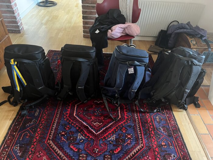 Fyra ryggsäckar står på en färgglad matta inomhus, redo för resa, omgivna av en stol och några klädesplagg.