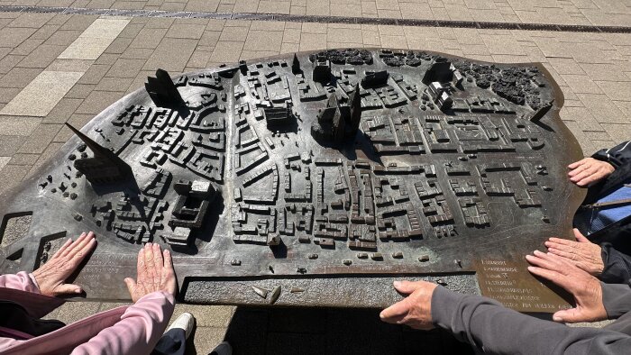Miniatyrmodell av en stadsplan i brons, omgiven av händer på en stenbelagd yta, med byggnader och gator tydligt markerade.