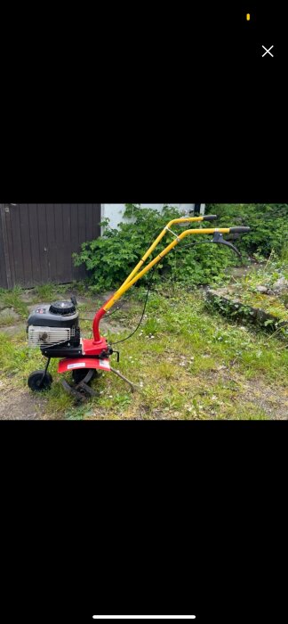 En röd och gul jordfräs med motor står på en gräsmatta nära en trädgård.