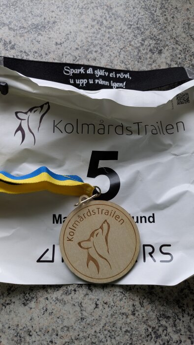 Startnummer med texten "KolmårdsTrailen", nummer 5 och en medalj med ett blågult band på en stenliknande bakgrund.