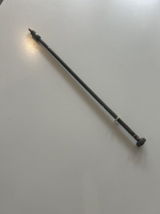 Lång metallstång med gängad del och skadad packning, från en stallkran, ligger på ett vitt bord.