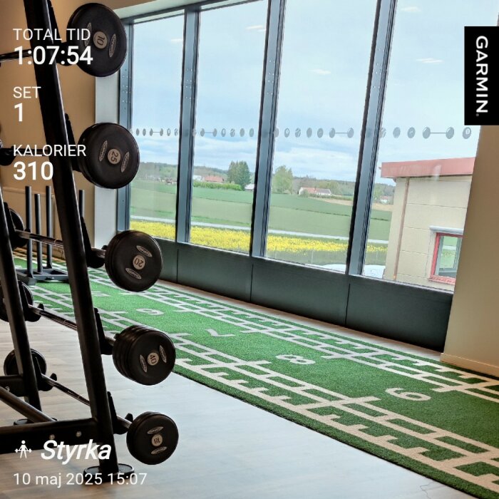 Gym med vikter, konstgräs för träning och stora fönster med utsikt över landskapet. Träningsinformation visas över bilden.