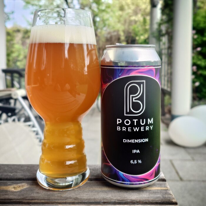 Ett glas fyllt med bärnstensfärgad öl bredvid en burk märkt Potum Brewery Dimension IPA, 6,5%, på ett bord utomhus.