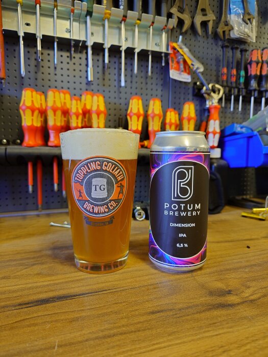 En ölglas med Toppling Goliath-logotyp bredvid en burk Potum Brewery Dimension IPA 6,5% på arbetsbänk med verktyg i bakgrunden.