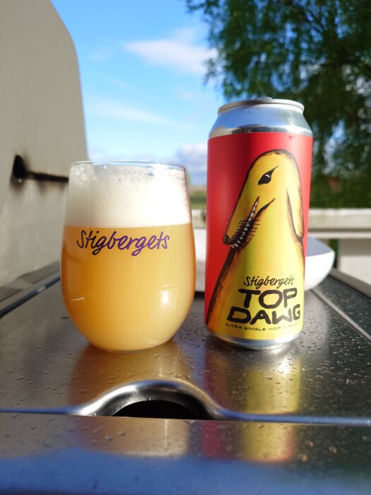 Burk och glas med öl från Stigbergets, etikett "Top Dawg", placerad på en grill med suddig bakgrund av en grönskande trädgård.