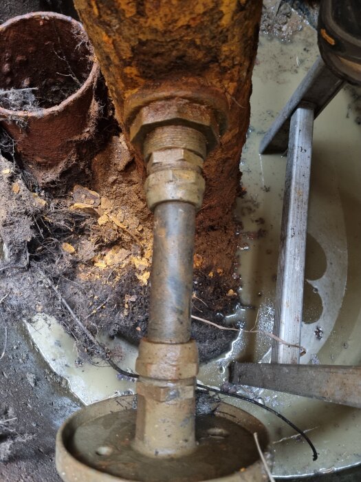 Rostig vattenpump i brunnhus med smuts och vatten runtomkring.