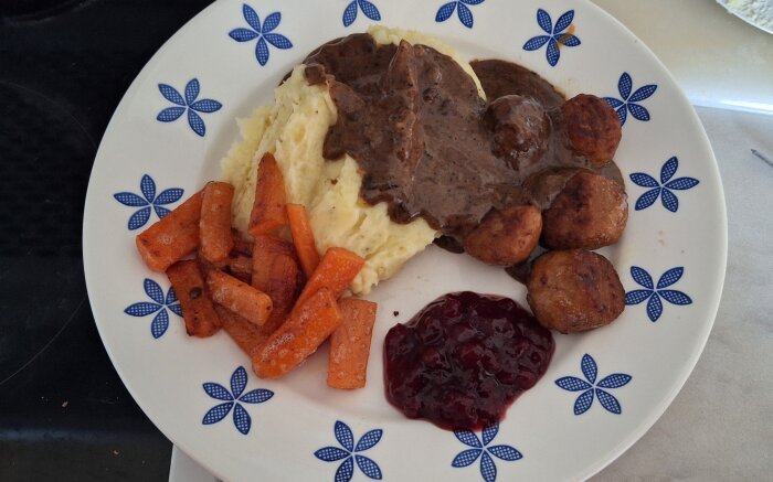 Köttbullar med potatismos, sås, stekta morötter och lingonsylt på en vit tallrik med blå mönster.