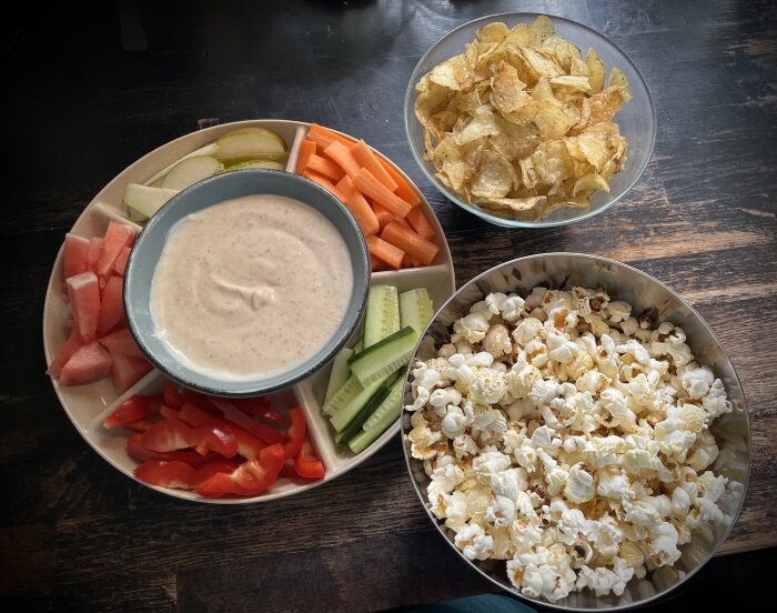 En tallrik med grönsaker och frukt samt en skål med dip, omgiven av chips och popcorn i två skålar.