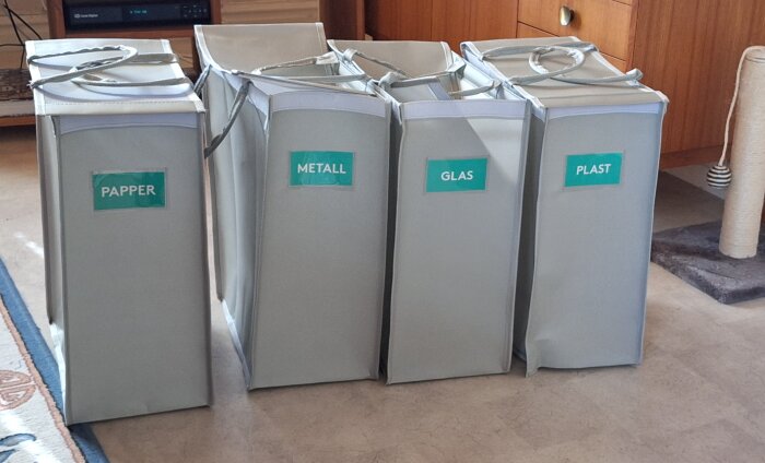 Fyra återvinningsbehållare för papper, metall, glas och plast står på rad i ett rum.