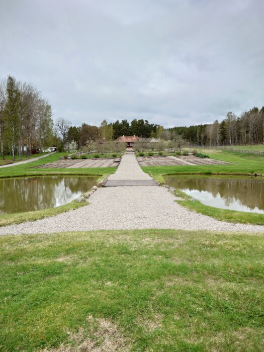 Grusgång leder fram till en röd villa omgiven av träd och gröna fält, med två symmetriska dammar på vardera sida om gången.