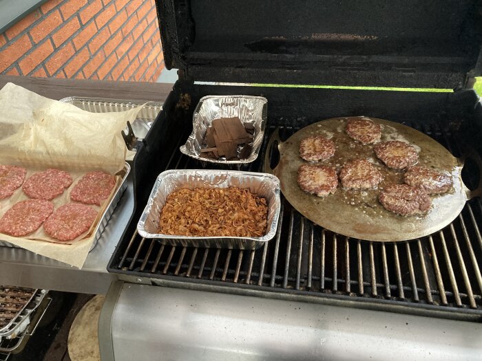 Grill med hamburgare på stekplatta, råa burgare på bakplåtspapper och karamelliserad lök i en aluminiumform.