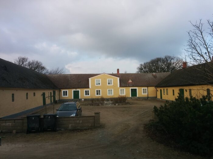 Gult gårdshus i gammal stil med innergård, omgiven av uthus. Två svarta soptunnor och en bil syns i förgrunden. Mulen himmel overhead.