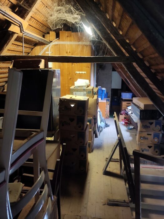 Ett gammalt vindsutrymme fyllt med kartonger, stolar, och andra föremål. Synliga takbjälkar och spindelväv i taket. Något mörk belysning.