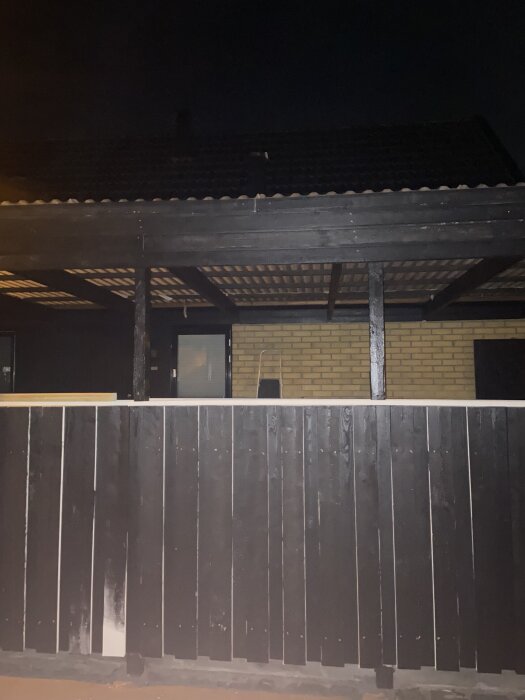 Carport på kvällen med svart trästaket och pergola, tegelvägg i bakgrunden, möjligt installationsområde för större glasruta från insidan.