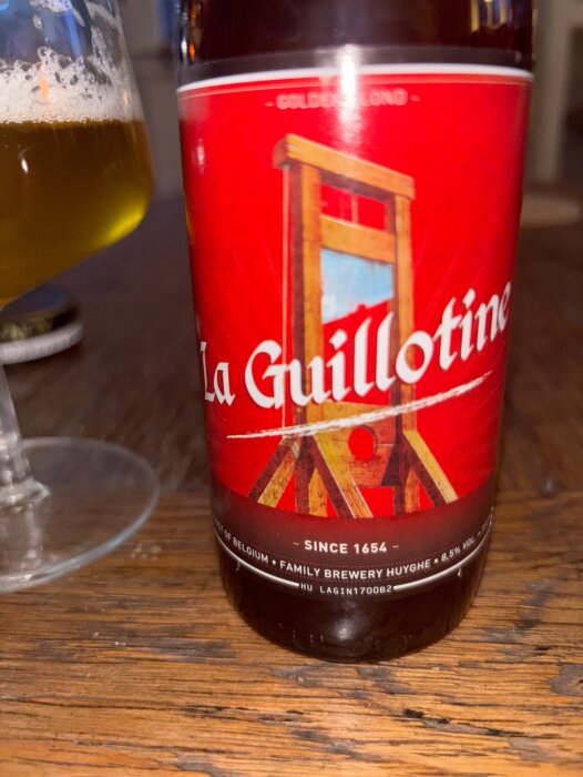 Ölflaska "La Guillotine" med ett glas öl, etikett visar en giljotin, traditionell belgisk brygd med citrus- och besksmak, Family Brewery Huyghe.