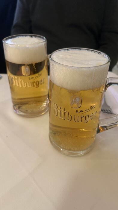 Två ölglas med Bitburger-öl på ett bord, med krämigt skum på toppen och en logotyp på glasen.