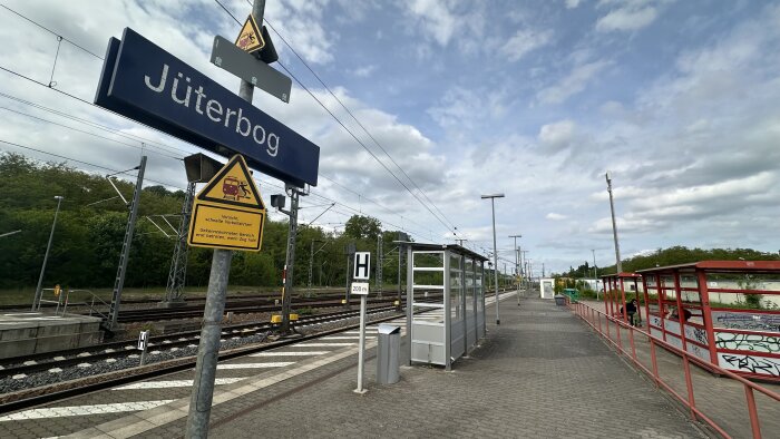 Jüterbog tågstation med skyltar, perrong och järnvägsspår under en molnig himmel. Kustskydd med graffiti syns till höger.