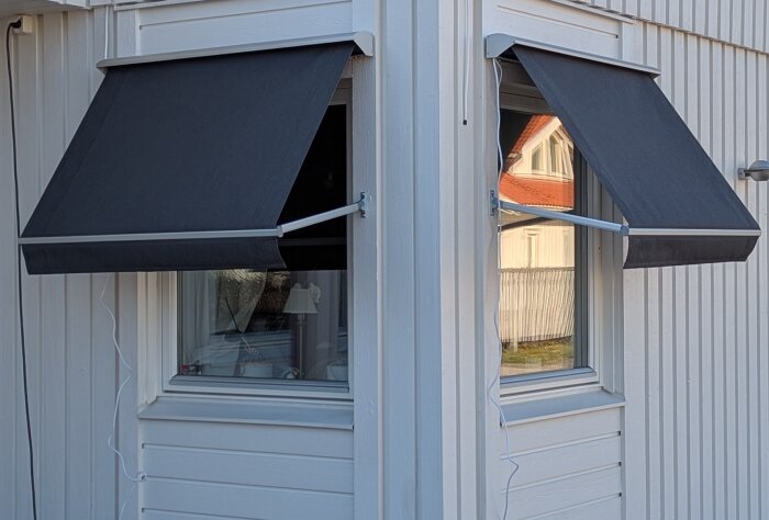 Grå markiser på två fönster av ett vitt hus.