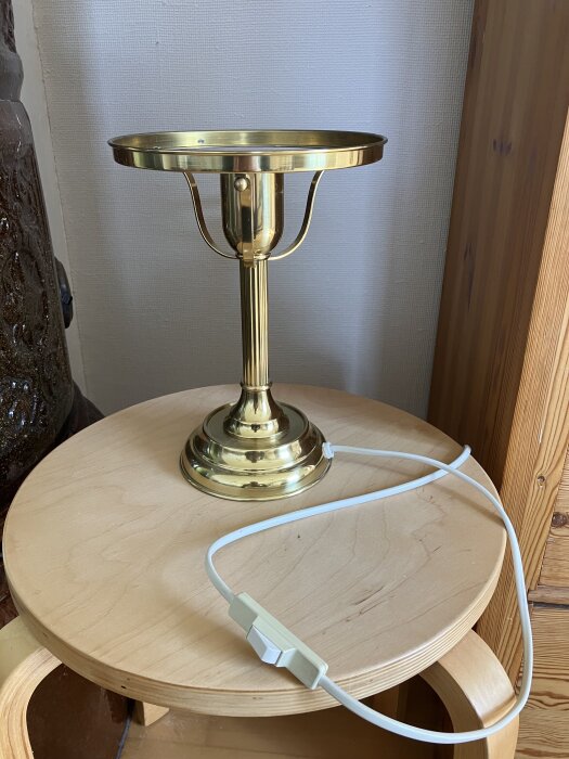 Guldfärgad halvgammal bordslampa utan lampskärm med en vit sladd och brytare, placerad på en rund träyta mot en vit vägg.