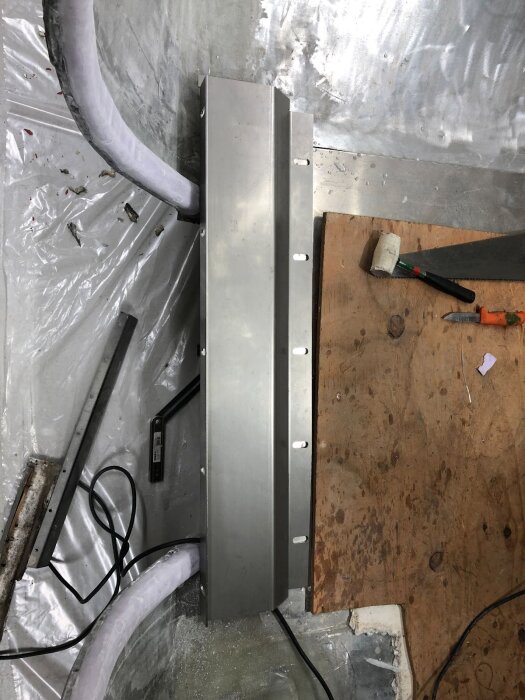 Rostfri plåt används som stark tröskel under dörrpost i byggprojekt, omgiven av träskiva och verktyg, med glasfiber och kablar i bakgrunden.
