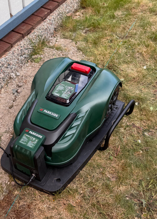 Grön robotgräsklippare av märket Parkside på laddstation vid gräsplätt nära en husvägg.