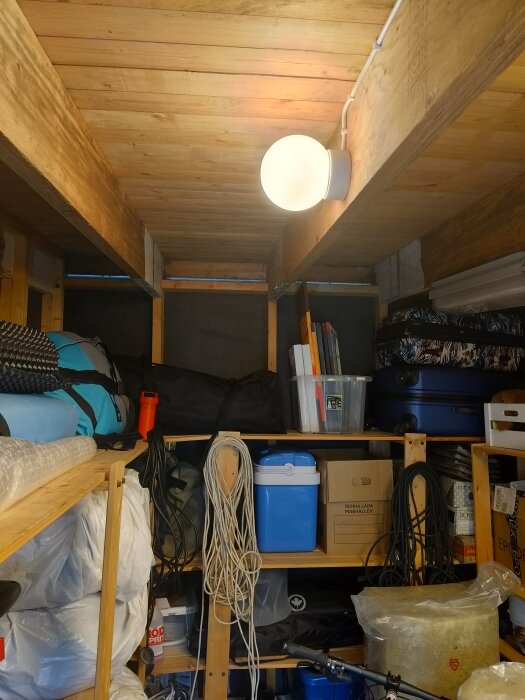 Förråd med trähyllor fyllda med föremål, en glödlampa i taket och synliga bjälkar.