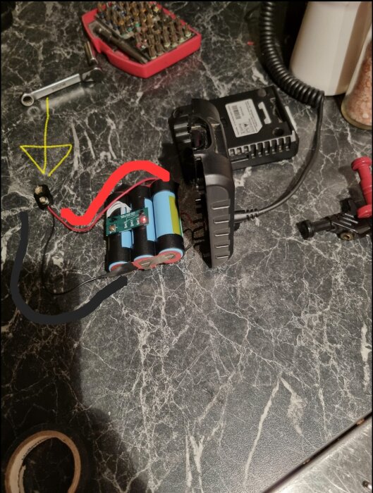 Bilden visar tre seriekopplade battericeller med en 9V-kontakt tejpad på, omgiven av verktyg. Gul, röd och svart markering för kontaktpunkter.