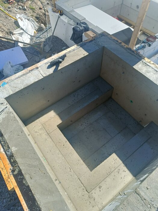 Betongbalja med bänkar på tre sidor, visar gropar och ojämnheter. Verktyg och material syns runt omkring på byggarbetsplatsen.