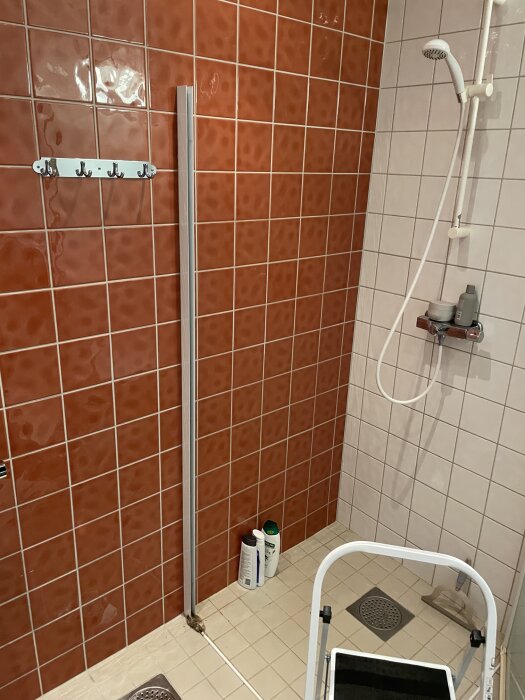 Duschhörn med borttagen vägg, röda och vita kakelväggar, silikontätad list och duscharmatur. Stegpall och flaskor på golvet.
