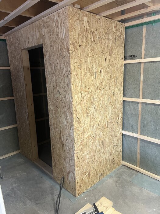 Byggd struktur i OSB-skivor, del av ett pågående byggprojekt med framtida toalett och gipstak i bakgrunden.