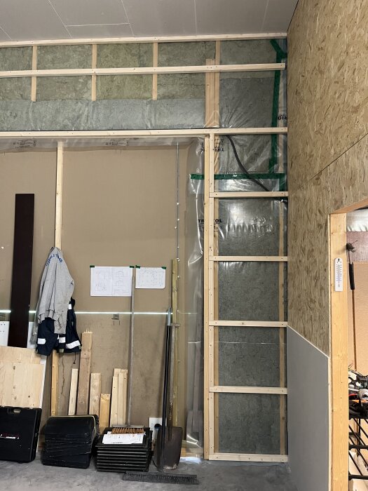 Bygget av en framtida toalett med regelverk, isolering och gipsskivor. Verktyg och byggmaterial ligger mot väggen. Planritning syns på väggen.