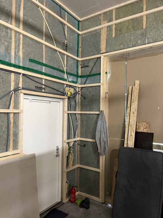 Väggar med trästommar och isolering, elektriska kablar dras. Vit dörr, brandsläckare, gipsskivor och byggmaterial syns i hörnet.