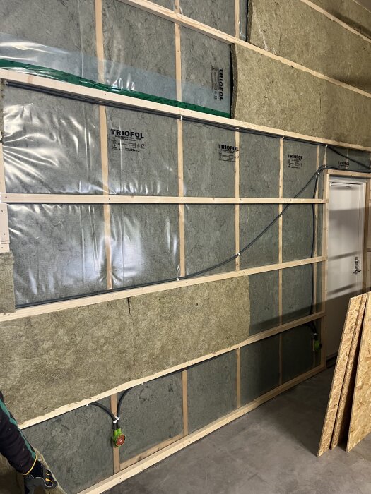 Väggkonstruktion med isolering av märket Triofol, samt ledningsdragning och en dörr som döljs delvis av OSB-skivor i förgrunden.
