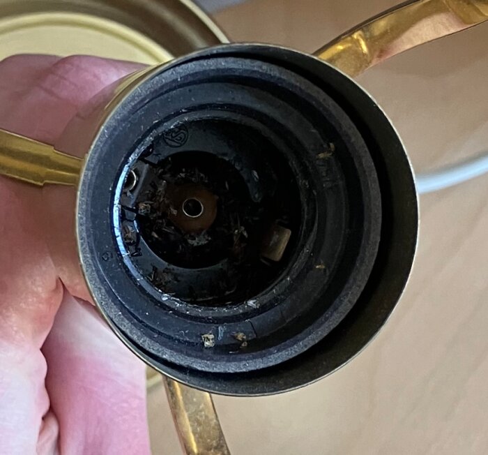 Lampinsats i närbild, med fokus på ett centralt hål där en glödlampa skulle skruvas i. Omgiven av guldfärgade detaljer.