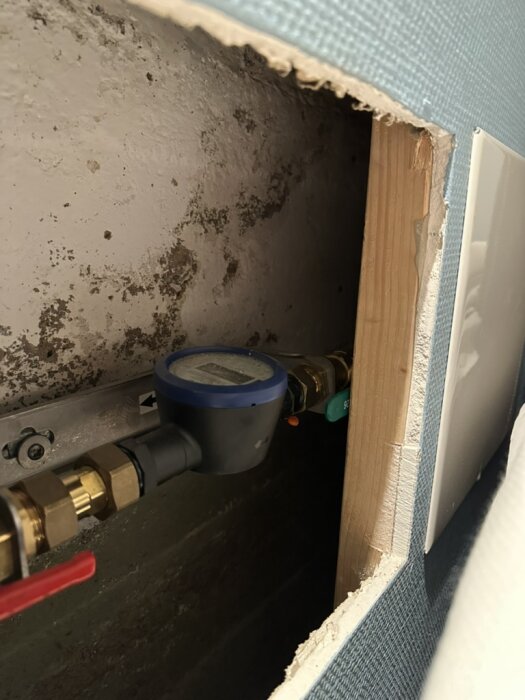 Väggutskärning i källare med synlig rörinstallation och fläckar på väggen; blå vävtapet runt öppningen.