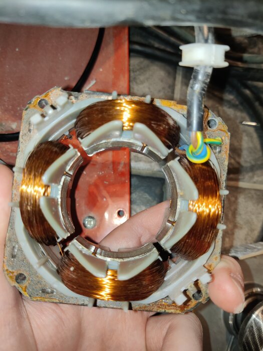 Närbild av statorlindningen i en elektrisk motor, med koppartråd virad runt en vit plastkärna, en del av en trasig byggfläkt.