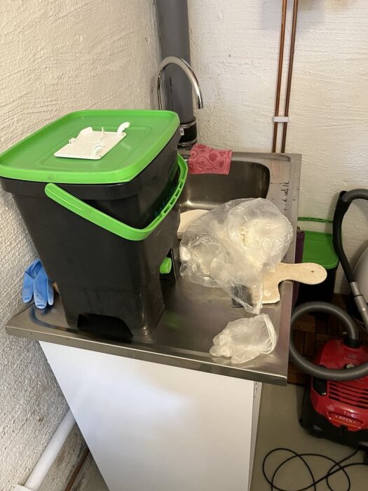 Diskbänk med grön hink, handfat och rör, plastpåsar och en dammsugare syns bredvid.