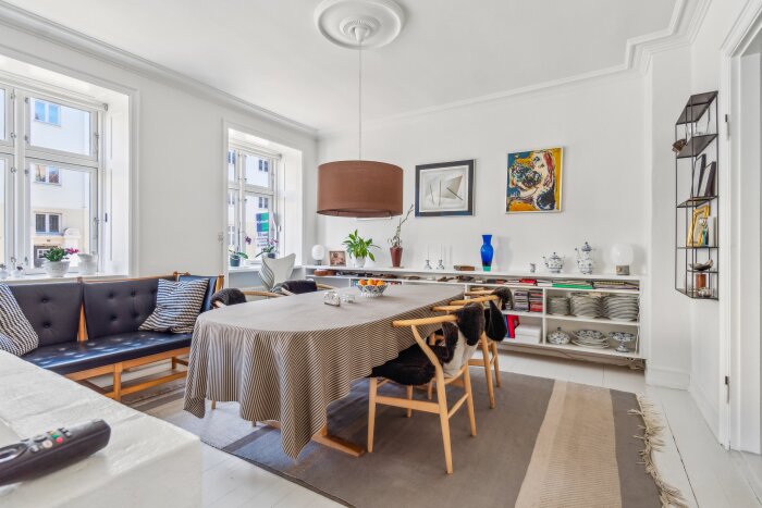 Ett ljust matsalsrum med ett stort bord täckt med randig duk, omgiven av stolar och fönster som släpper in mycket dagsljus.