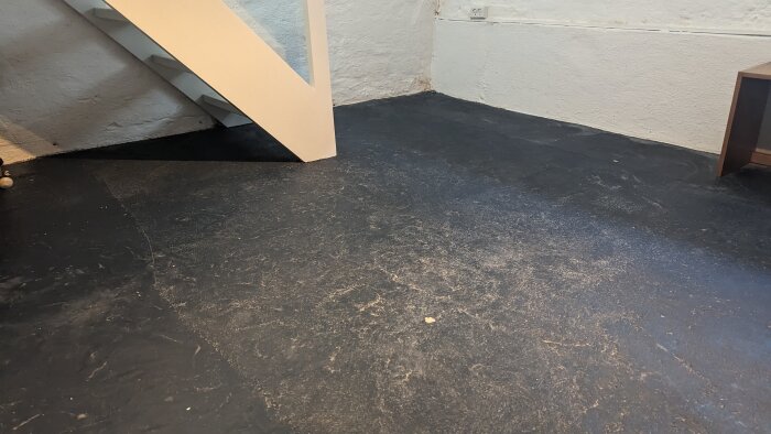 Oinredd källare med mörkt golv, vitmålade väggar och en trappa i hörnet.