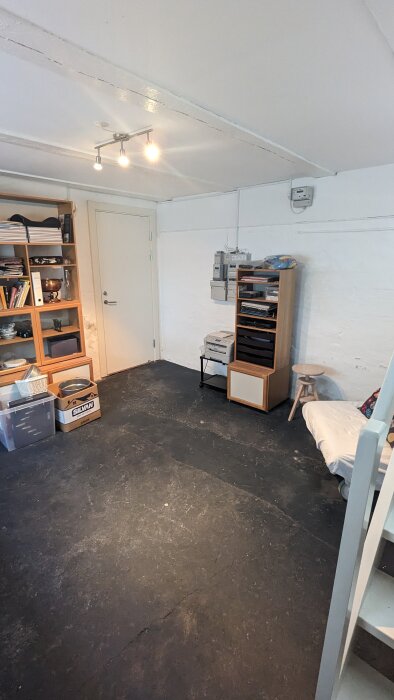 Oinredd källare med mörkt golv, bokhyllor fyllda med lådor och pärmar, samt en pall och soffa vid sidan.