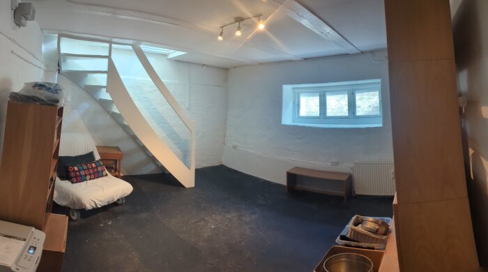 Oinredd källare med vitmålade väggar, mörkt golv, en trappa, en fåtölj med kudde och ett fönster med ljusinsläpp.