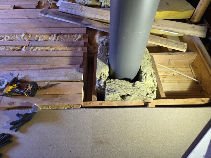 Golvspånskivor och trägolv runt ett stort grått kaminrör med isolering. Byggverktyg och arbetshandskar ligger på golvet.