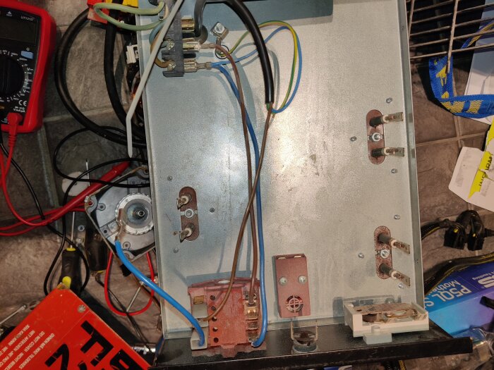 Öppen elektrisk kopplingsbox med lösa kablar, instrument och verktyg. En röd multimeter syns på vänster sida.